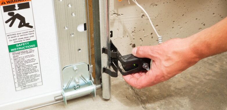 A hand adjusts a garage door sensor mounted near the floor in an FL home. A warning label and safety instructions are visible on the door, and wiring is attached to the garage doors Fort Meyers sensor bracket.