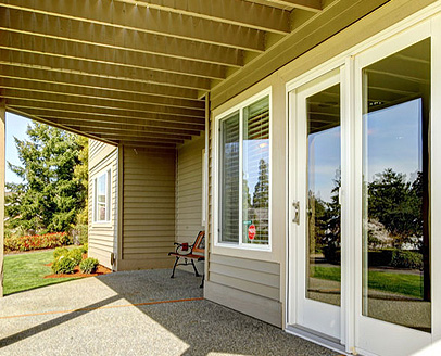 A covered patio area with beige siding, glass doors, a window with blinds, a small bench, and a view of a green lawn and trees in the background—ideal for relaxing after exploring FL or updating your garage doors Fort Meyers style.