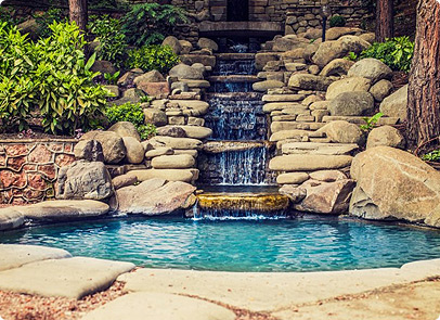 A small, clear pool surrounded by large rocks with a cascading waterfall flowing down stone steps, lush green plants, and trees in the background—an oasis perfect for relaxing after choosing garage doors in Fort Myers, FL.