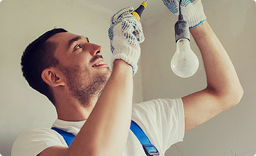 A man wearing work gloves and overalls is installing or replacing a light bulb on a ceiling, using a screwdriver and smiling as he works—dedicated to quality repairs just like with garage doors in Fort Meyers, FL.