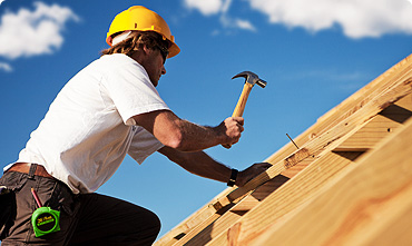 A construction worker in FL wearing a yellow hard hat and white shirt uses a hammer to drive a nail into a wooden roof frame under a blue sky. A green tape measure is clipped to his belt, while nearby garage doors Fort Meyers are being installed.