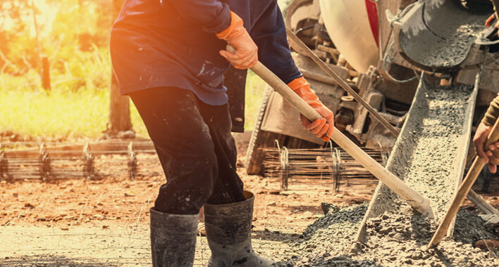 A person wearing rubber boots and gloves spreads wet concrete from a mixer truck at a construction site, preparing the foundation for new garage doors in Fort Myers, FL, with sunlight streaming in the background.