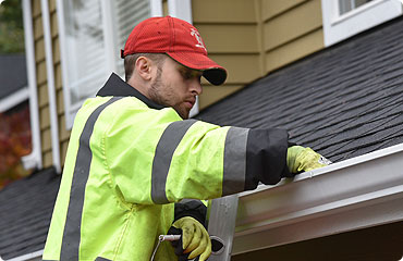 A person in a red cap and high-visibility jacket cleans debris from a house gutter while standing on a ladder next to a yellow siding building near garage doors in Fort Meyers, FL.