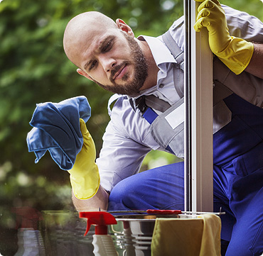 A man wearing yellow rubber gloves and a work uniform is cleaning a window with a blue cloth. Cleaning spray bottles are visible on the windowsill, with greenery outside—a spotless scene typical of FL homes or garage doors Fort Meyers services.