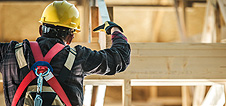 A construction worker in FL, wearing a yellow hard hat, safety harness, and gloves, works on a wooden structure near garage doors Fort Meyers, using a saw indoors as sunlight streams in.