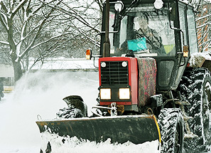 A red snowplow tractor clears snow from a road on a winter day in FL, with trees and parked cars—some near garage doors—visible in the background as the plow pushes snow to the side.